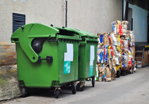 A large green communal rubbish bin with a rounded lid and black handle, positioned on an urban pavement next to a textured grey wall with a brick section at the base. The bin has two white reflective strips on its front and is set on small black wheels at the bottom, indicating it is mobile for waste collection. To the right of the bin, there are several tightly packed bales of flattened cardboard boxes in various colors, including brown, blue, red, and white, stacked in a slightly leaning formation against the building. The scene appears to be in a town or city area, consistent with drainage and lighting typical of an inland UK locality, potentially within Victoria. The surroundings suggest a commercial or residential waste disposal context, as managed by Garden Clearance Victoria for rubbish clearance or waste collection services. The background includes part of a concrete wall, a small vent, and a building with large windows, all on a paved surface, indicative of an accessible urban environment for rubbish collection.
