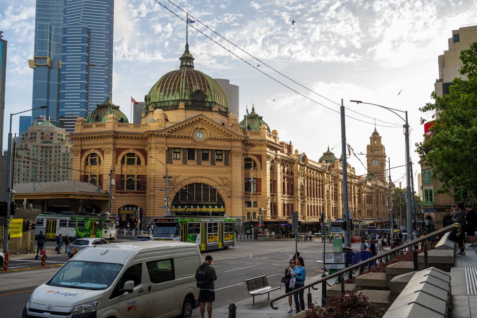A nighttime cityscape of Melbourne featuring illuminated skyscrapers, including the Eureka Tower with its distinctive spire, and historic buildings with pointed spires and domed rooftops. Bright stree